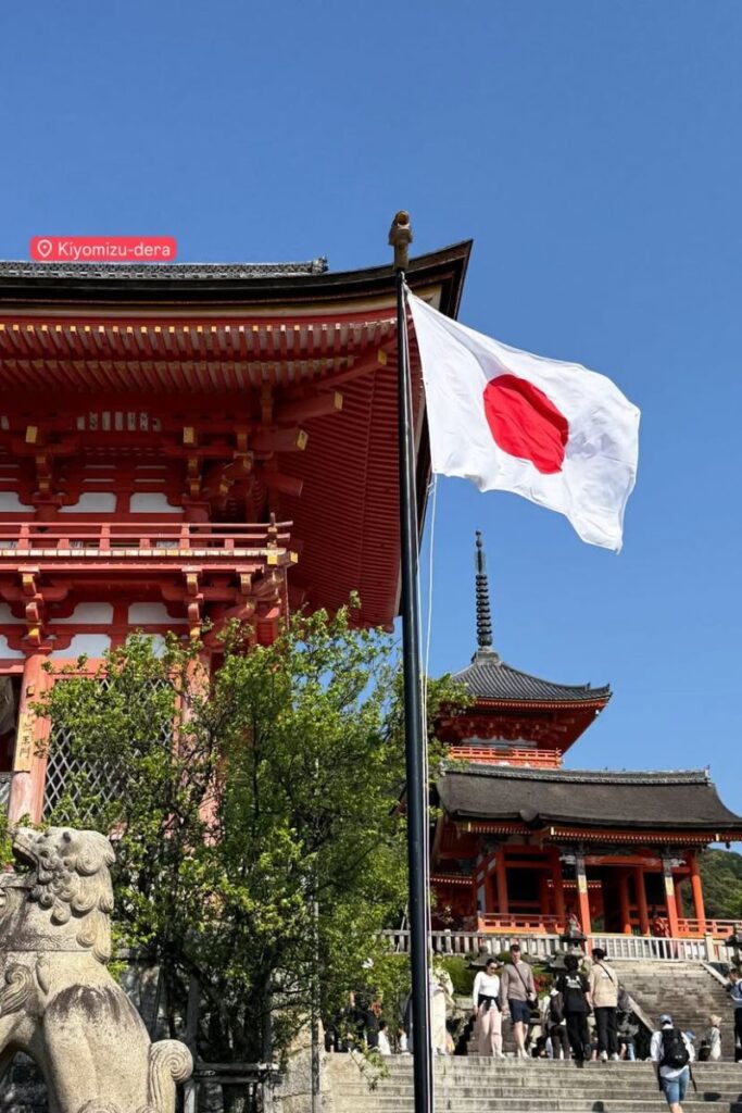 A Japanese flag waving in front of Kiyomizu-dera Temple in Kyoto, Japan, with traditional architecture in the background. Perfect for content on solo female travel in Japan, best place for solo female travel, or traveling alone in Japan.