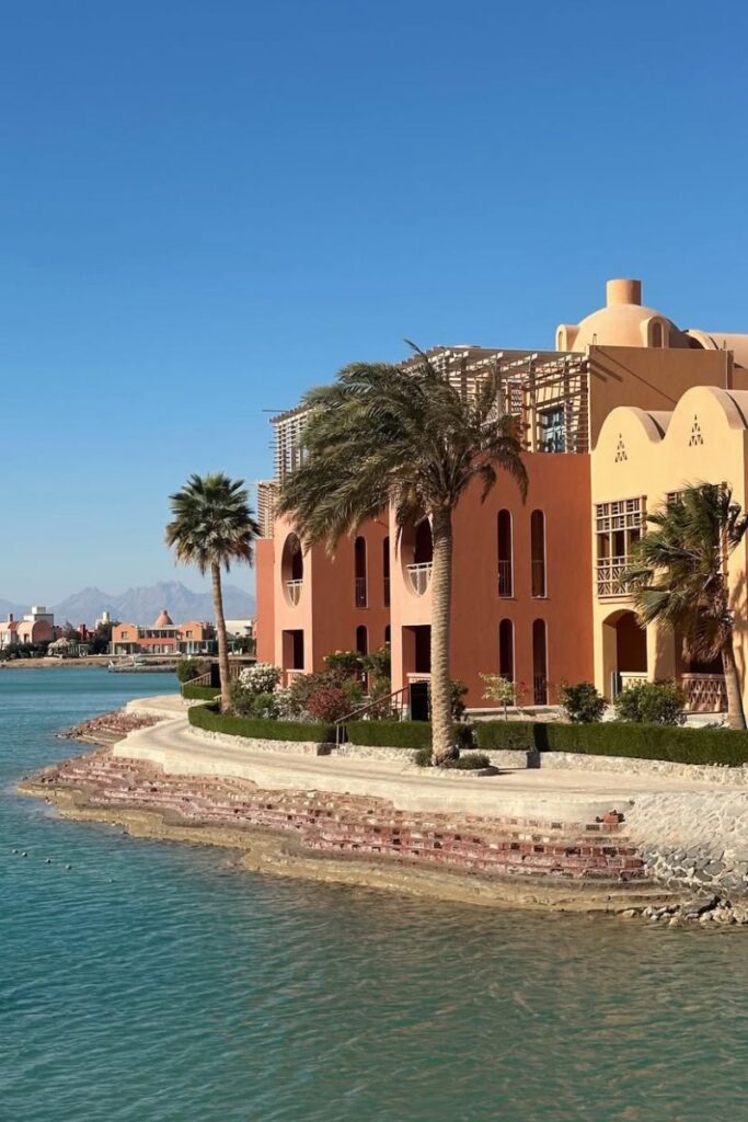 A terracotta-colored hotel sits beside a quiet lagoon, with palm trees blowing in the wind and distant mountain peaks. ideal for exploring whether to stay in Hurghada or El Gouna and the best hotels in Hurghada for snorkeling.