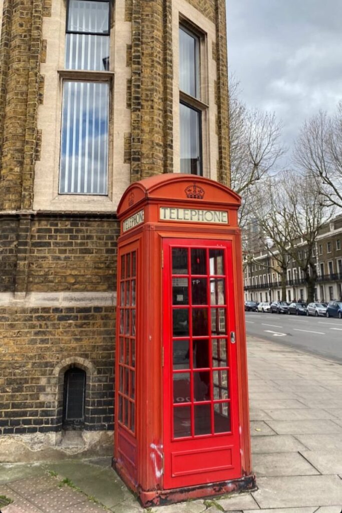 A classic red telephone booth in London, positioned on a quiet sidewalk next to a historic brick building. Great for content on traveling to London alone, best cities for solo female travel, or first time traveling alone.
