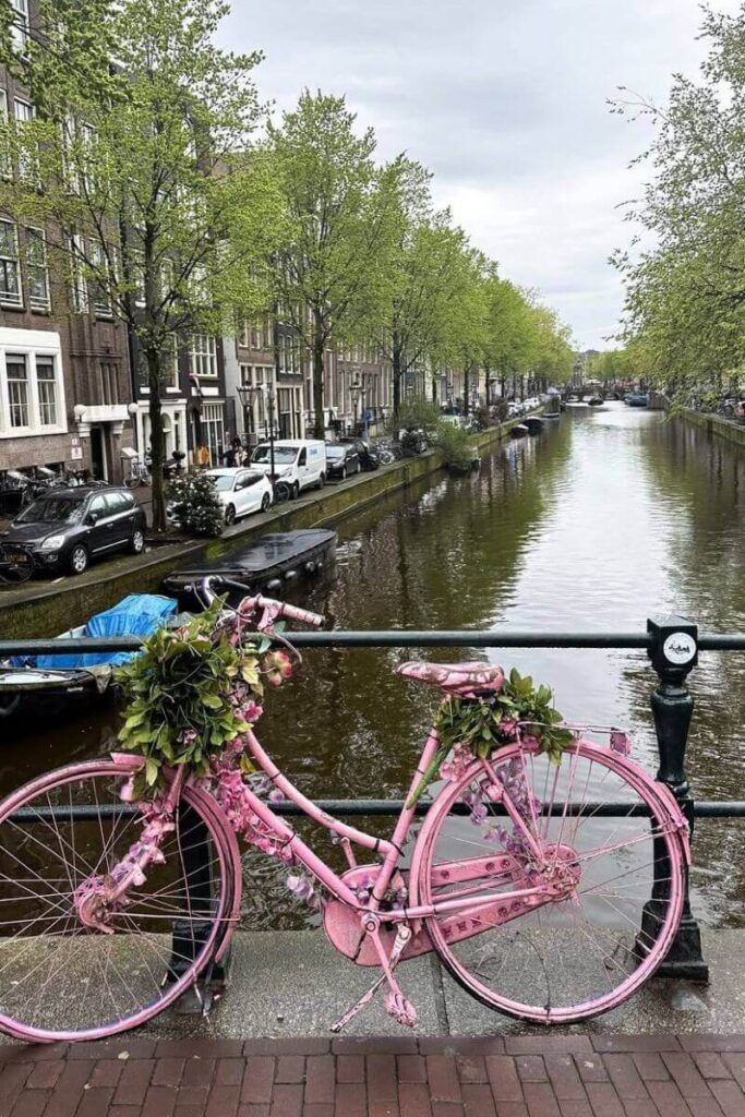 A charming canal in Amsterdam lined with trees and houseboats, with a bright pink bicycle decorated with flowers on a bridge railing. A perfect feature for posts on Europe solo travel, best places in Europe to travel solo, or best cities to travel solo in Europe.
