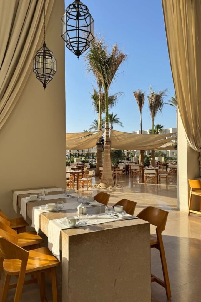 Stylish open-air restaurant with beige curtains, geometric hanging lanterns, and rows of tables facing a sunny garden filled with palm trees. This inviting breakfast space belongs to one of the best hotels in Hurghada Egypt all inclusive, ideal for relaxing mornings at a 5-star hotel in Hurghada Egypt.