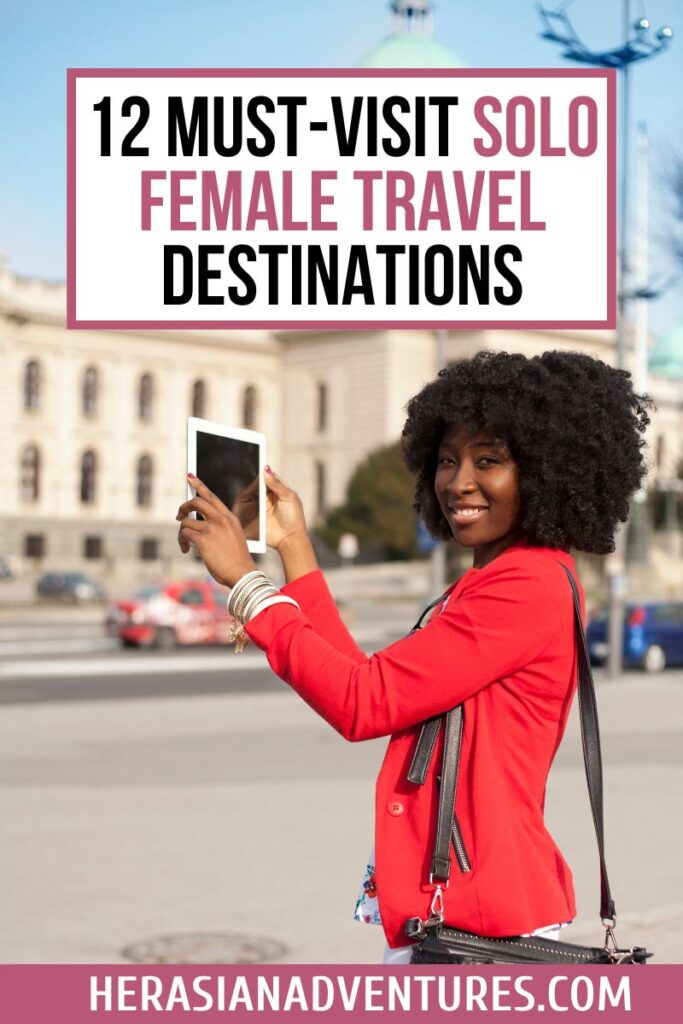 A smiling woman in a red jacket holding a tablet, posing confidently in front of a European-style building, with the overlay text: "12 MUST-VISIT SOLO FEMALE TRAVEL DESTINATIONS." Great for guides on solo female travel destinations or best places to travel for solo female travelers.