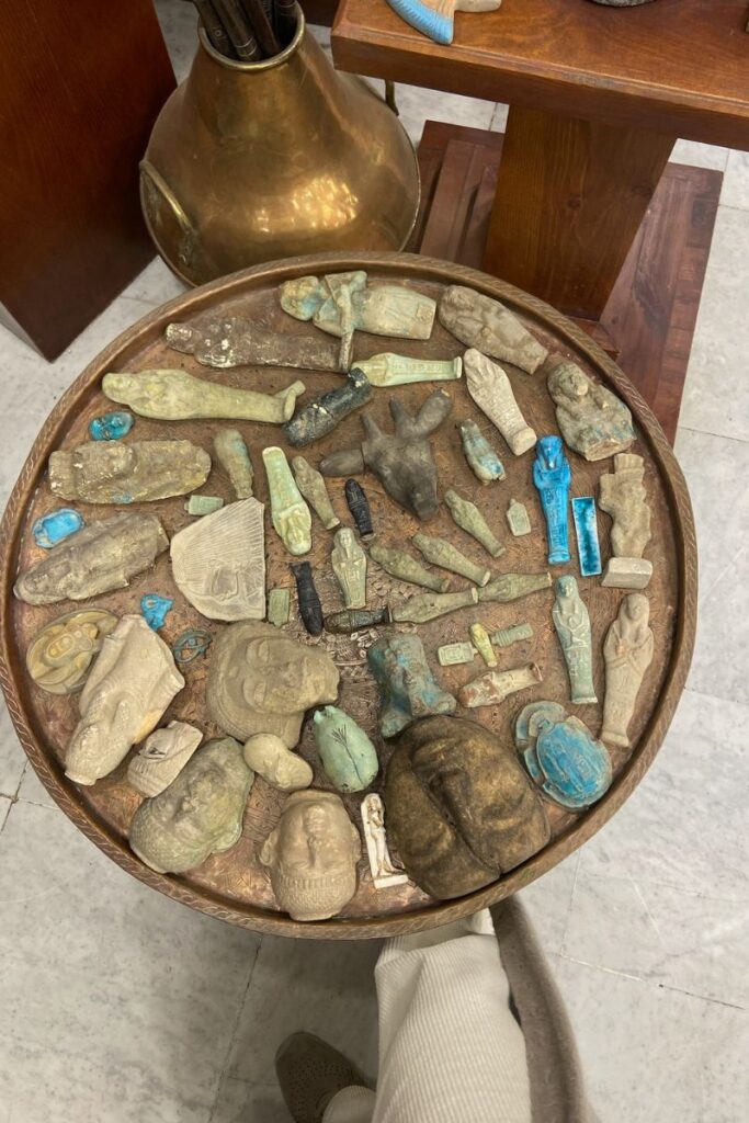 A round tray filled with an eclectic mix of ancient-style Egyptian artifacts, including shabti figurines, faience amulets, and broken headpieces in various colors. These replicas are iconic Egyptian souvenirs that reflect the country's Pharaonic history and are ideal gifts from Egypt. This is a great representation of what to buy from Egypt and must-have souvenirs in Egypt for history buffs.