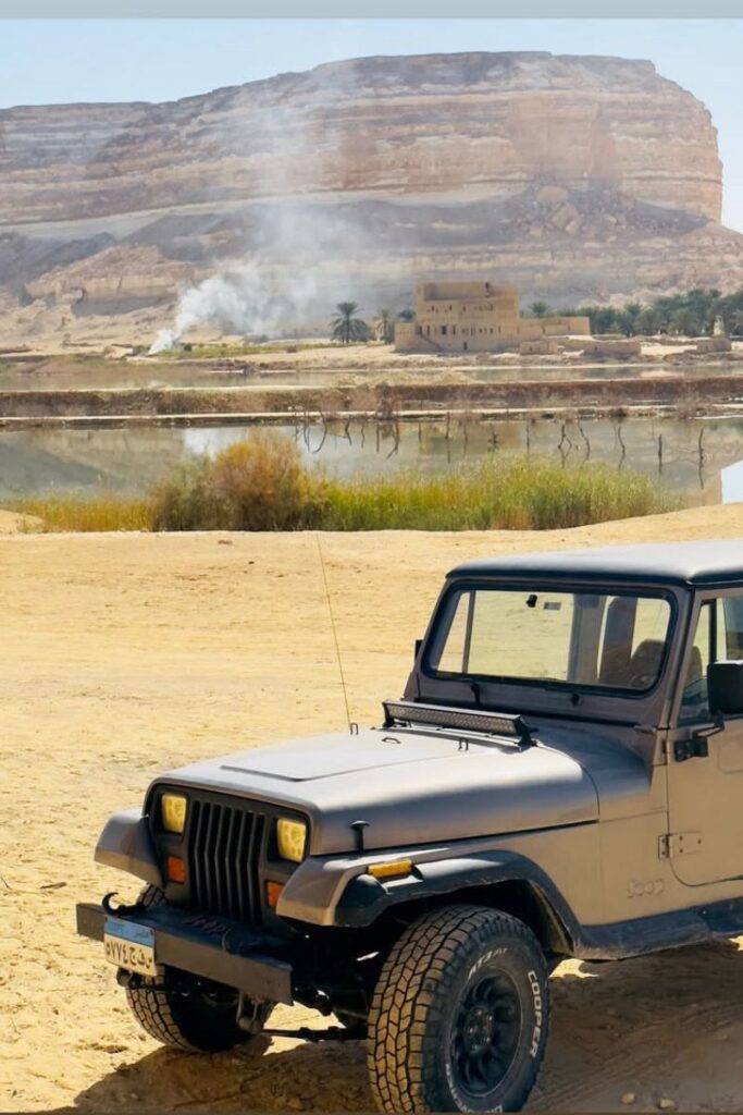 A black 4x4 Jeep parked on sandy terrain near a reflective lake and desert cliffs, with smoke rising in the distance—an adventurous scene along the cairo to siwa route through the egyptian oasis.