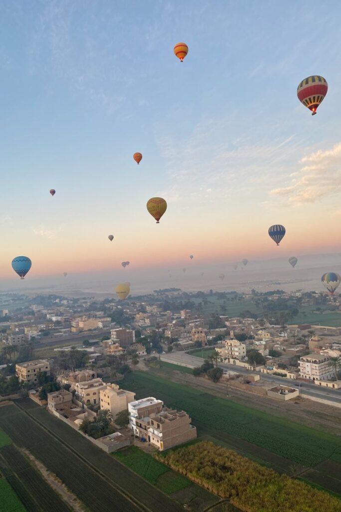 Dozens of colorful hot air balloons float over green fields and the town of Luxor at sunrise. A magical experience for solo female travelers in Egypt in one of the safest places to travel in Egypt.