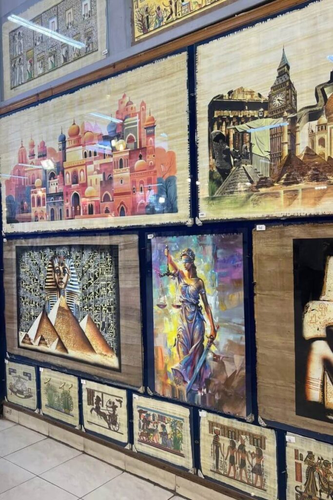 A display of framed papyrus artwork featuring Egyptian pyramids, deities, Lady Justice, and European landmarks, merging ancient and modern themes. These pieces are standout Egyptian souvenirs that appeal to a wide range of tastes and are among the best things to buy in Egypt. Great for tourists looking for unique Egypt gifts and visual keepsakes from their trip.