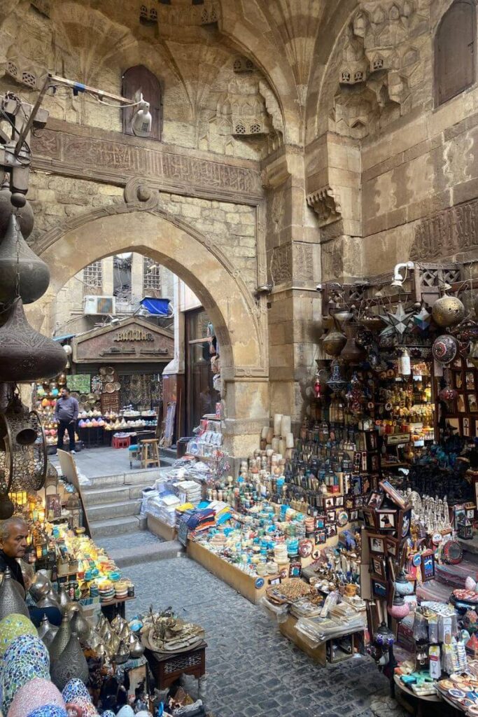 A packed alleyway market under a historic archway in Cairo, filled with lanterns, perfumes, ceramics, spices, and textiles. This vibrant setting is a top destination for Egypt shopping and a hotspot for finding good souvenirs from Egypt. From traditional crafts to modern items, it's ideal for anyone wondering what to buy from Egypt and where to find authentic Egyptian gifts.