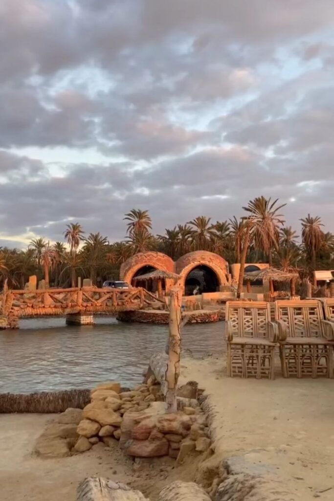Rustic lodge-style buildings with palm-thatched roofs line a lakefront in Siwa Oasis, Egypt, with bamboo chairs and dramatic skies overhead—ideal for showcasing unique hotels in Siwa Oasis or a Siwa Oasis hotel experience.
