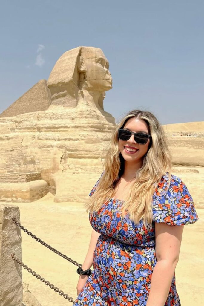 Solo female traveler smiles in front of the Great Sphinx of Giza on a sunny day, wearing sunglasses and a floral dress. An iconic site for women asking is it safe to travel to Egypt and exploring Cairo’s historic wonders.
