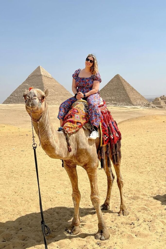 A solo female traveler smiles while riding a decorated camel in front of the Pyramids of Giza under a clear blue sky. A classic Egypt travel moment for women wondering is Egypt safe for solo female travelers and seeking adventure while traveling in Cairo.