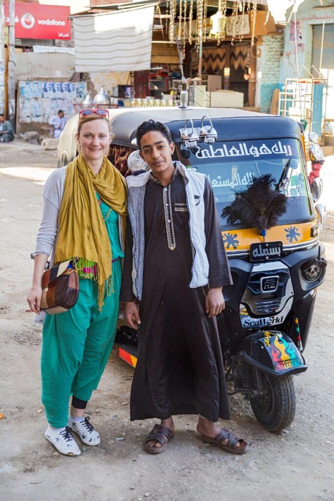 A solo female traveler poses with a local man in traditional dress beside a brightly decorated tuk-tuk in an Egyptian town. A candid scene showing how to stay safe when visiting Egypt and that Egypt is safe for women with local awareness.