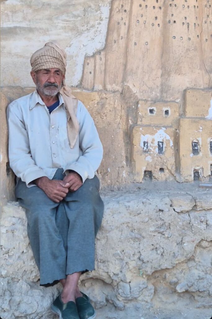 An elderly Egyptian man wearing traditional clothes sits peacefully against a rustic wall painted with village motifs. A quiet cultural snapshot appreciated by women travelers in Egypt seeking authentic experiences.
