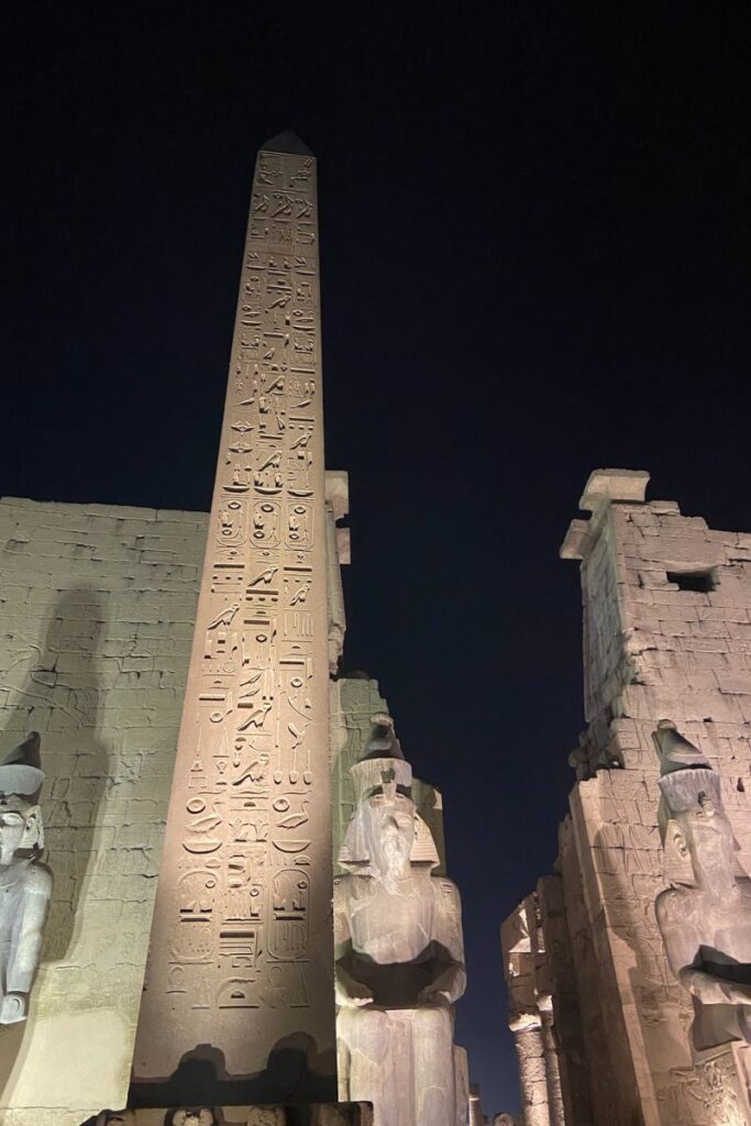 Tall obelisk covered in hieroglyphics flanked by colossal seated pharaoh statues at Luxor Temple, a must-see Egyptian temple known for its grandeur and historical importance.
