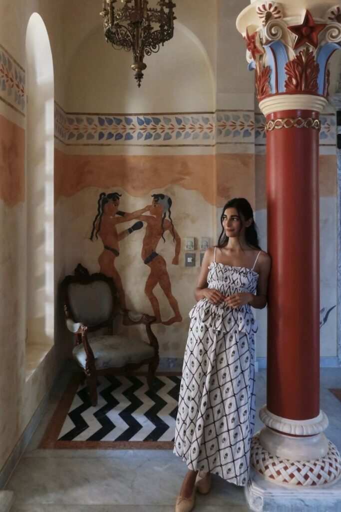 Woman in a printed two-piece outfit stands beside a red pillar in a richly decorated hotel alcove with a Minoan-style fresco of two women boxing on the wall. This charming and artistic corner reflects the kind of thoughtful design seen in boutique hotels in Hurghada Egypt and is ideal for travelers looking for the best hotel to stay in Hurghada Egypt.
