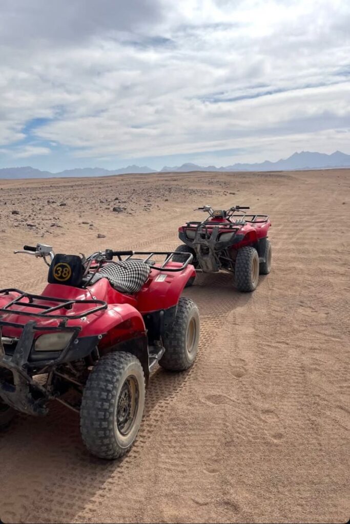Two red quad bikes parked on a sandy desert trail with mountains in the distance under a partly cloudy sky. Perfect for a buggy tour Hurghada and one of the top fun things to do in Hurghada.