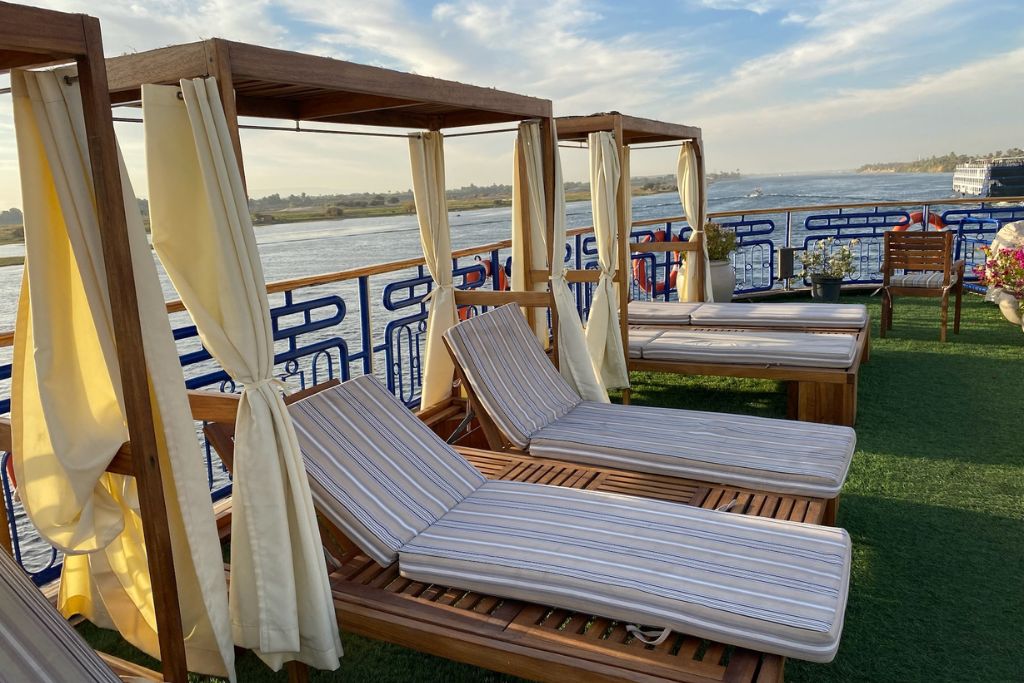 Comfortable wooden sun loungers and canopy beds overlook the Nile River from the deck of a cruise ship. A relaxing option for Americans traveling to Egypt and a peaceful experience for women traveling solo in Egypt.