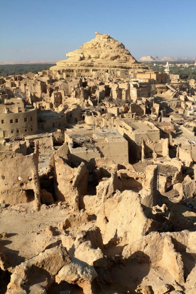 Wide view of the crumbling Shali Fortress ruins in the center of Siwa Oasis Egypt, with traditional mud-brick buildings set against the backdrop of a beautiful desert and palm-dotted horizon.