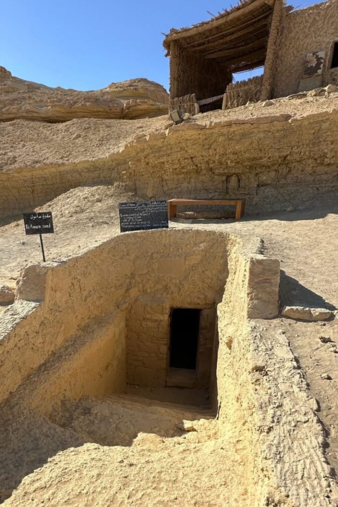 Entrance to an ancient tomb in the desert with signs reading “Si Amun tomb,” featuring handwritten Arabic and English descriptions—offering a glimpse into the archaeological wonders in the oasis of Siwa, Egypt.