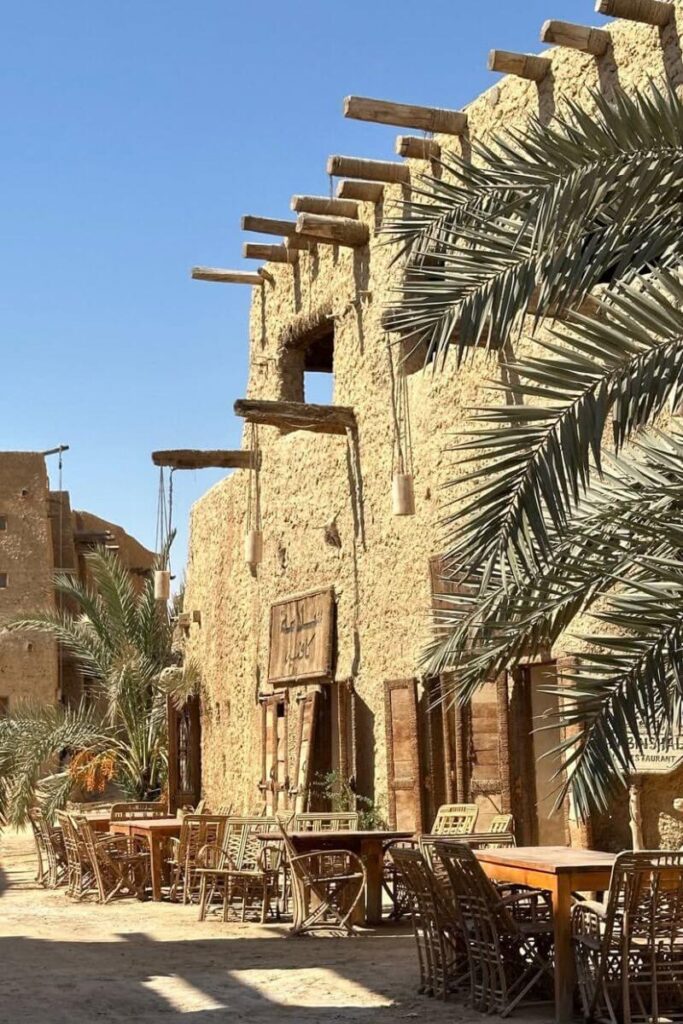 Outdoor dining setup along a sandy path beside a palm tree and traditional mudbrick building in Siwa Egypt, with a wooden sign written in Arabic—ideal for showcasing hotel in Siwa and unique Siwa Oasis hotels.