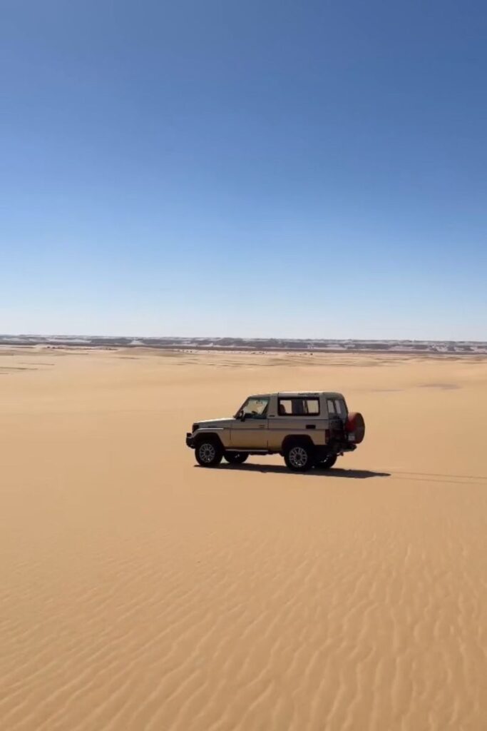 A 4x4 vehicle parked alone in a vast sea of golden sand dunes in Siwa Oasis, under a bright blue sky—symbolizing the freedom of desert exploration and the thrill of a Siwa Oasis trip.