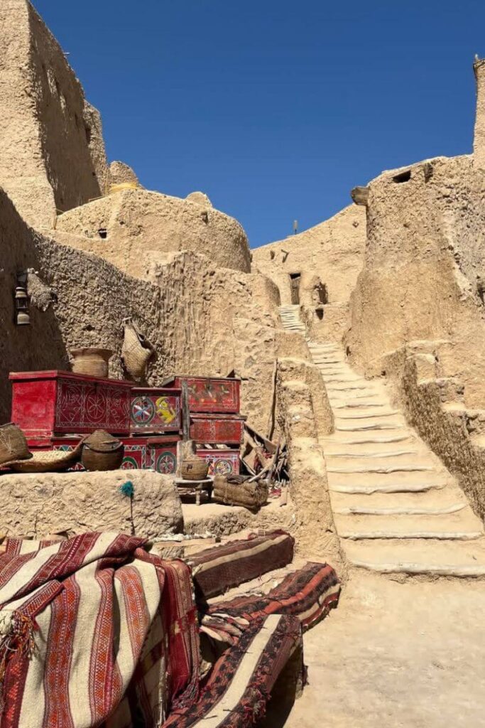Rustic stairway carved into the desert walls, with vibrant woven rugs and painted red chests in the foreground, capturing the authentic feel of the oasis of siwa and nearby hotels in siwa egypt.