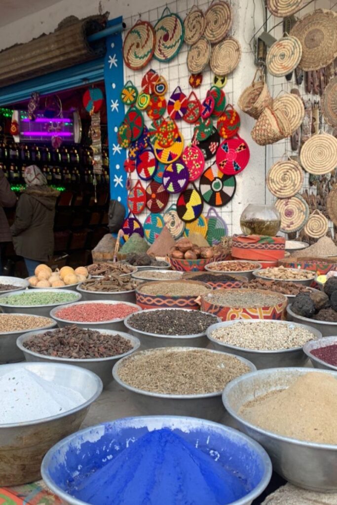 A colorful spice market in Egypt showing metal bowls filled with spices and herbs in all colors, in front of woven baskets hanging on the wall. This lively scene is perfect for travelers interested in what to buy in Egypt, especially for authentic, aromatic souvenirs from Egypt like spices, henna, or incense. A top spot when Egypt shopping for gifts that awaken the senses.