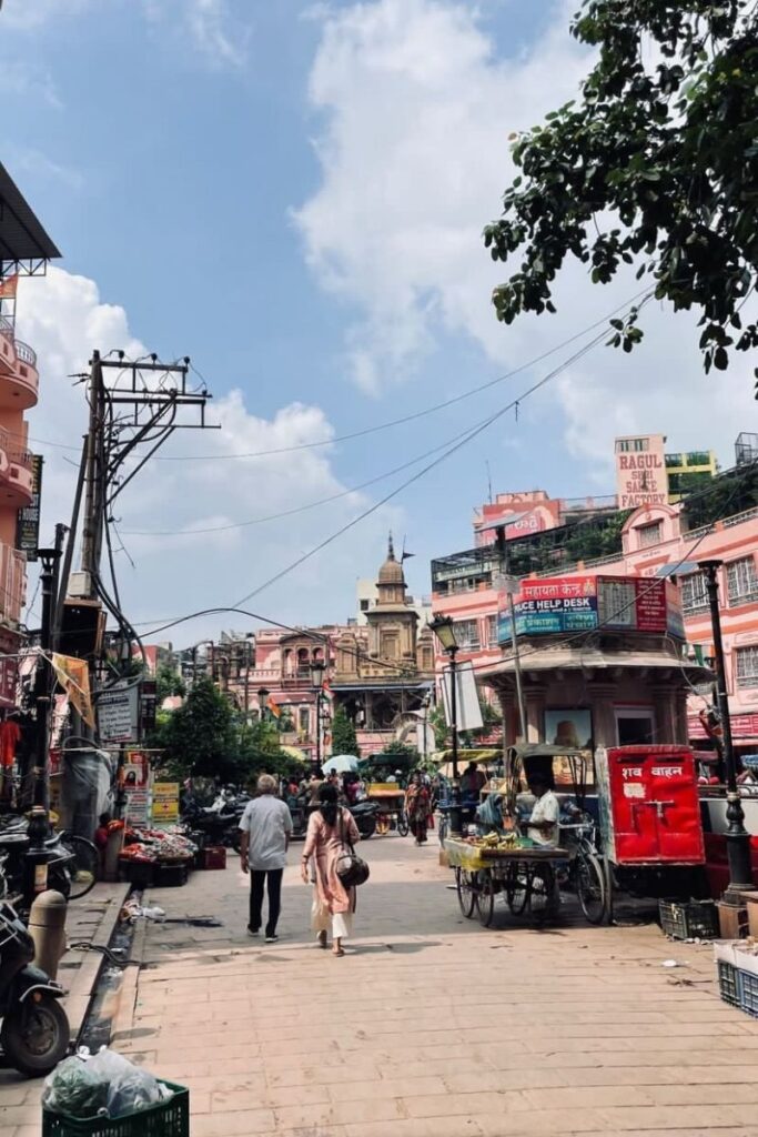 A bustling street in Varanasi, India, filled with pink-hued buildings, street vendors, and locals walking beneath a bright sky—an authentic experience for solo female travelers in India. Great for content on best solo travel destinations India and places for female solo travel in India.