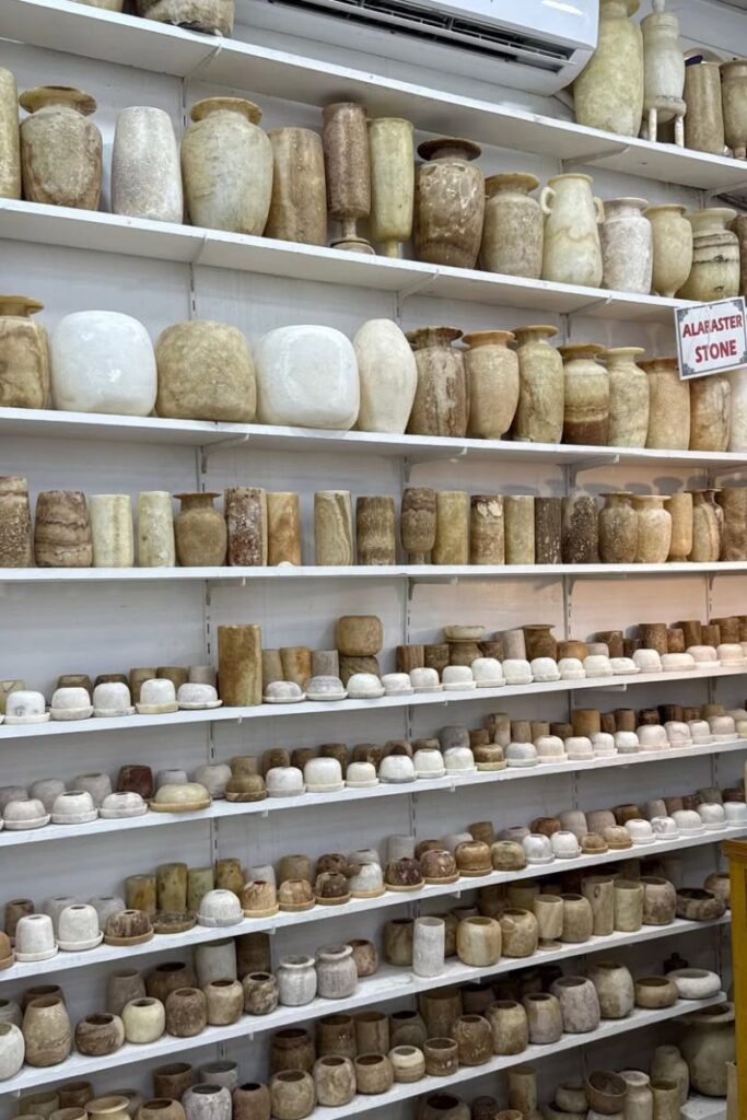 Shelves filled with hand-carved alabaster vases, jars, and bowls in creamy beige and white tones, sold in an Egyptian shop under a sign labeled “Alabaster Stone.” These unique stone pieces are elegant Egyptian souvenirs and top suggestions for things to buy from Egypt that blend beauty, history, and craftsmanship. Ideal gifts from Egypt that stand out.