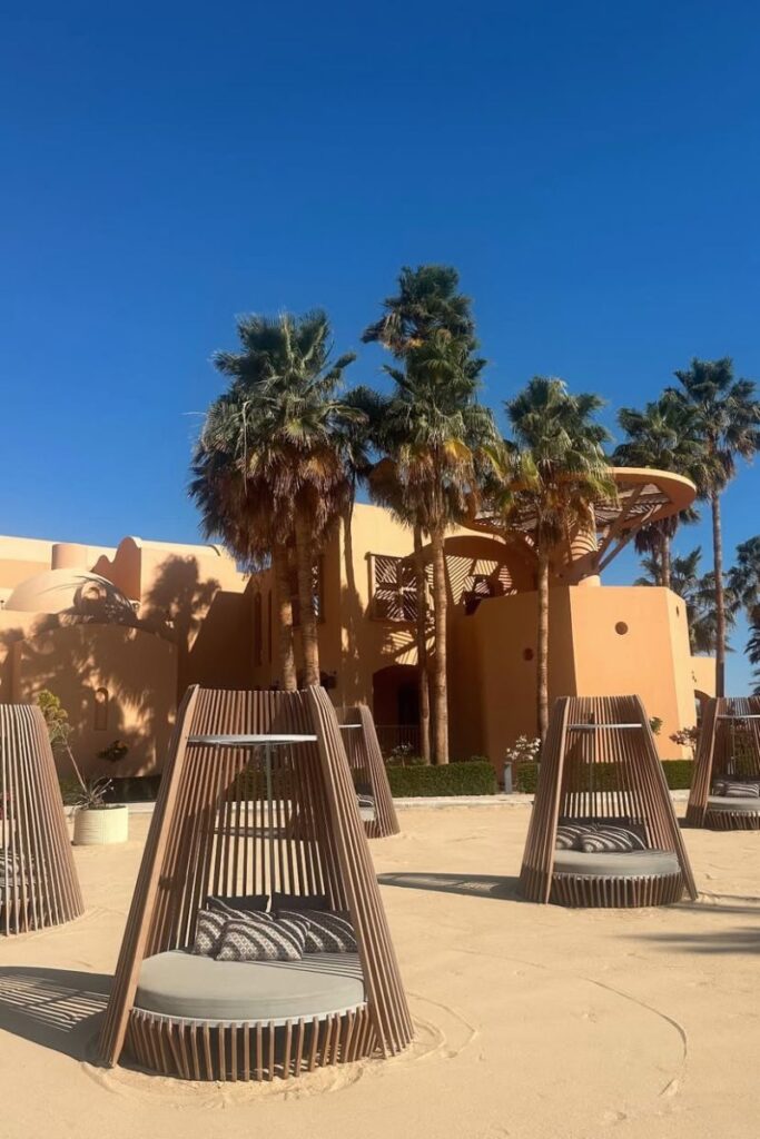 Modern wooden beach pods with cushions are arranged on sandy ground in front of a warm-toned resort surrounded by palm trees. Perfect for best aqua park hotels in Hurghada and stylish resorts for couples or singles.