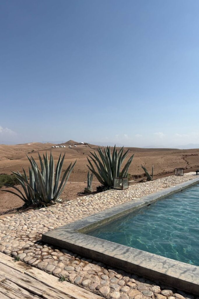 A luxurious pool lined with smooth stone sits in a quiet desert landscape with tall agave plants, under a clear blue sky. This remote escape is one of the best warm destinations in October for travelers looking to relax in nature with warm, dry weather.