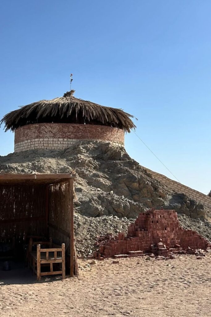 A traditional circular stone and thatch hut perched on a rocky hill in a desert village near Hurghada, with handmade seating nearby. Visiting local Bedouin communities is one of the most authentic things to do in Hurghada Red Sea Egypt and an enriching addition to day trips from Hurghada