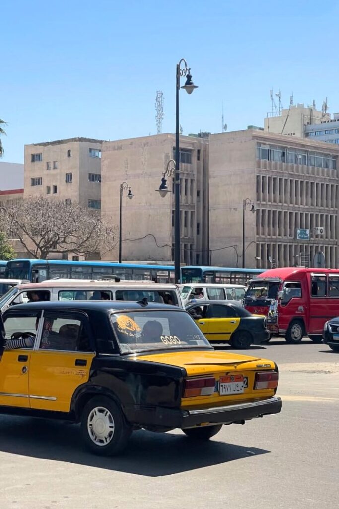 Busy urban street in downtown Cairo with yellow and black taxis, red and white minibuses, and concrete government buildings in the background. Navigating this energetic capital is a key cultural experience when traveling to Cairo from Hurghada and exploring Hurghada travel guide extensions.