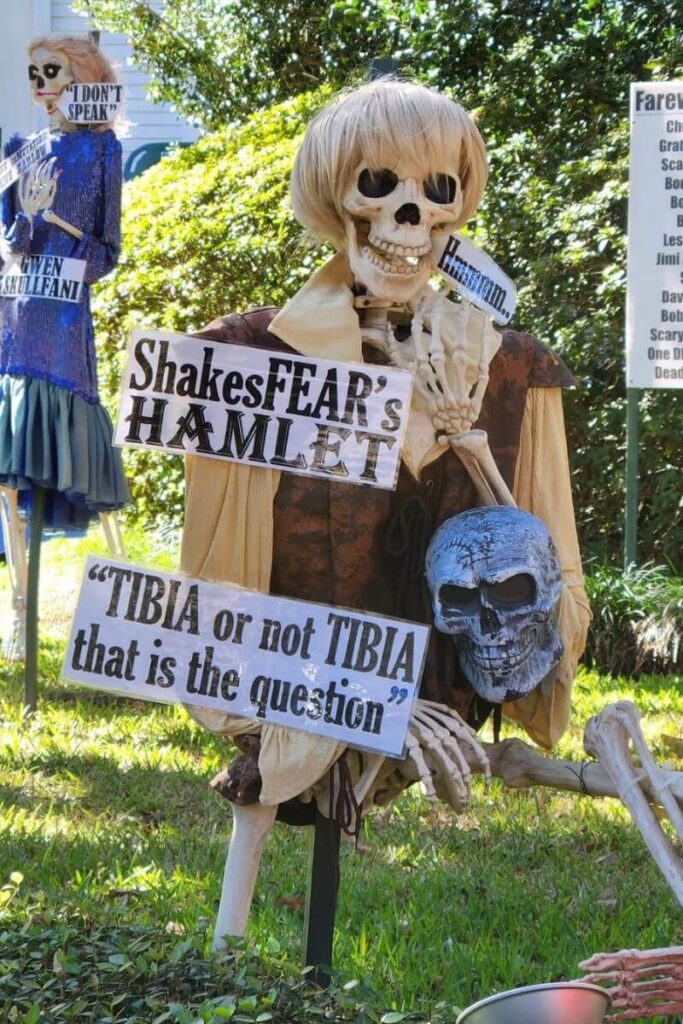 A skeleton dressed as Shakespeare’s Hamlet holds a skull with a sign reading “Tibia or not Tibia that is the question,” a playful decoration at one of the best places to visit for Halloween lovers.