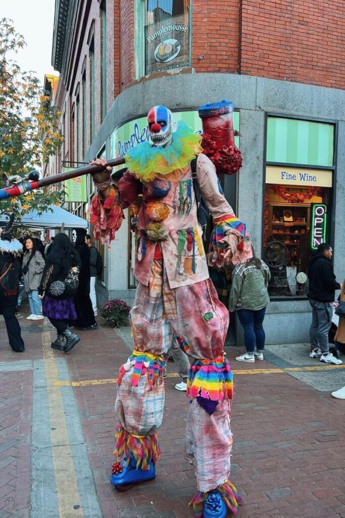 A tall, scary clown in tattered rainbow clothing and holding a prop weapon entertains onlookers, a highlight of spooky places to visit for Halloween in the US.