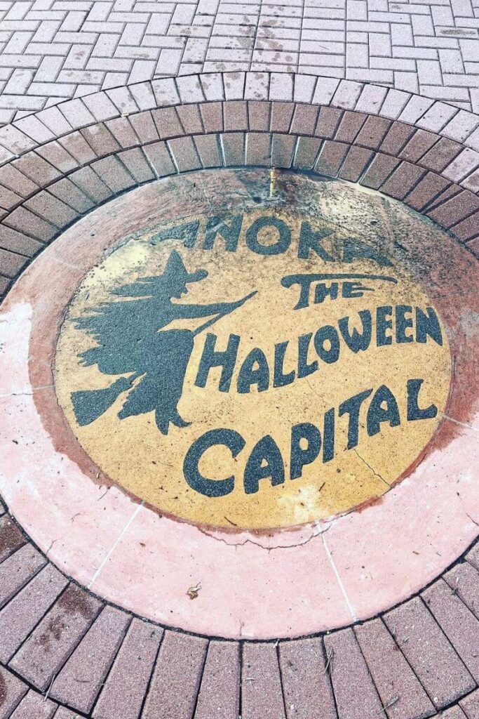 A round sidewalk plaque in Anoka, Minnesota, reading “The Halloween Capital” with a witch silhouette, marking one of the most famous Halloween towns in the US.