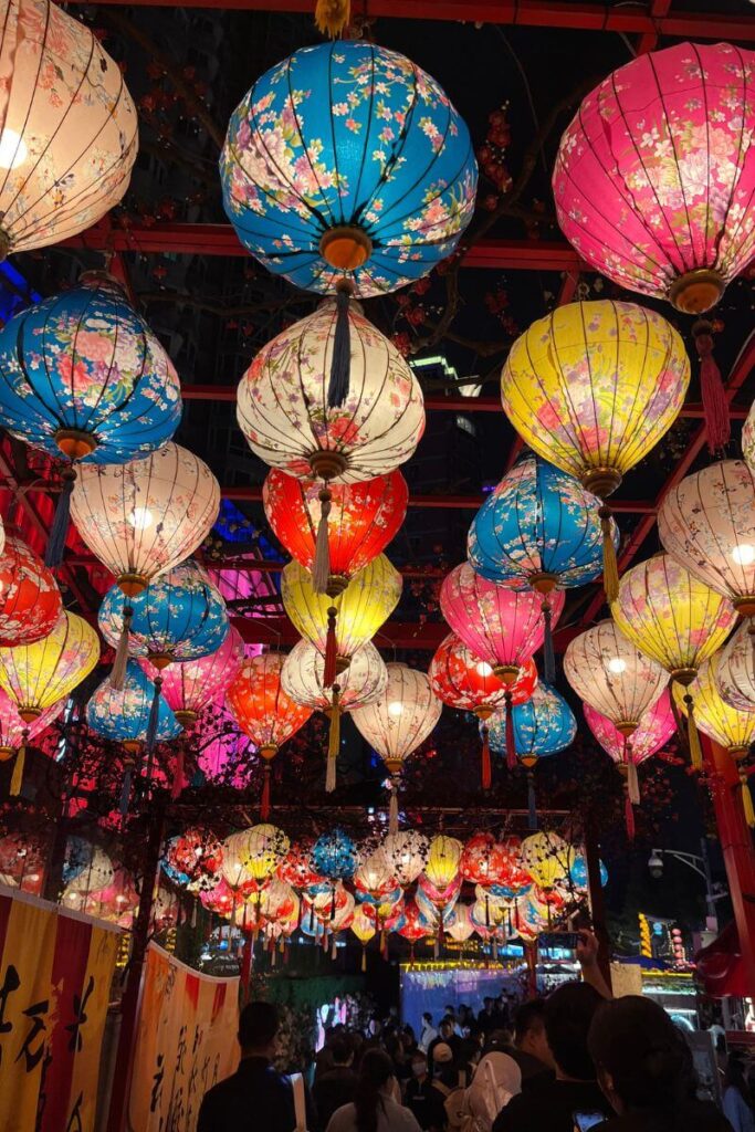 A festive night market glows with colorful hanging lanterns patterned with flowers, drawing crowds underneath. This cultural event is ideal for those planning a vacation in October and looking for warm cities with lively festivals and unique experiences.