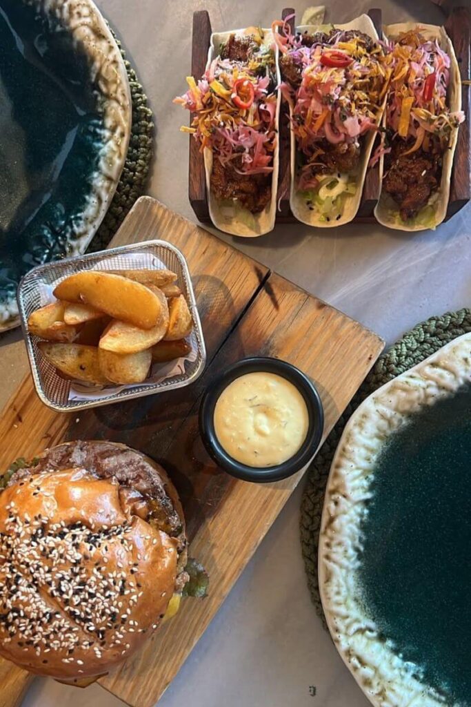 A close-up of a juicy sesame seed burger, golden potato wedges, creamy dipping sauce, and colorful tacos on a wooden serving board. Sampling international and fusion cuisine is one of the things to do in Hurghada town and a hidden gem in the Hurghada travel guide for food lovers.