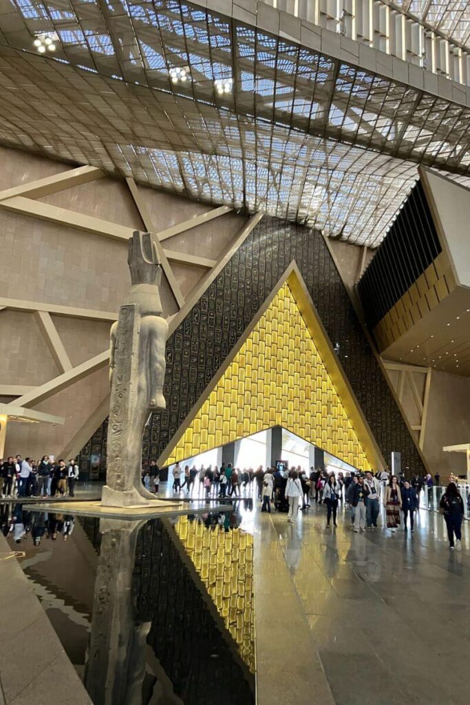 Interior of the Grand Egyptian Museum with visitors walking beneath a massive golden pyramid-shaped structure and ancient statue. A central experience on any Egypt itinerary 10 days or comprehensive Cairo Egypt itinerary.