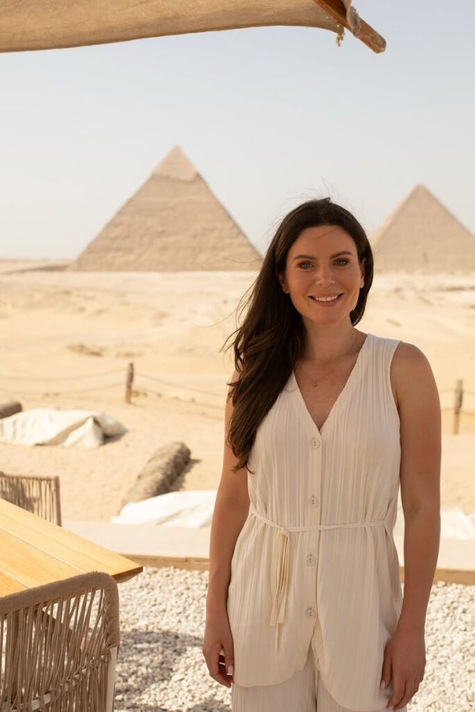 Smiling woman stands at a shaded outdoor café with the three Giza pyramids clearly visible behind her. A perfect shot from a 10 day Egypt itinerary or one week in Egypt itinerary exploring Giza and Cairo.