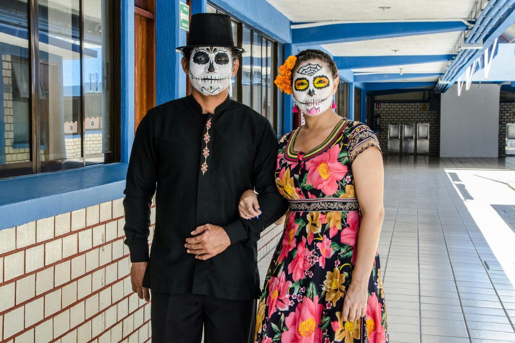 A couple in traditional Mexican attire with sugar skull face paint walks arm in arm, representing Día de los Muertos, a vibrant Halloween travel destination in Mexico.