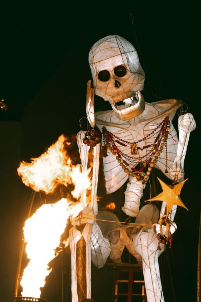 A towering illuminated skeleton puppet adorned with beads and holding a golden star is surrounded by bursts of fire during a nighttime festival, one of the best places to visit for Halloween lovers looking for dramatic outdoor celebrations.
