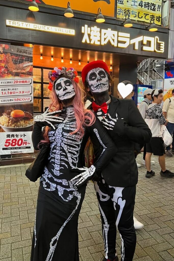 Two people in detailed skeleton costumes with face paint, floral crowns, and formalwear pose for photos in a busy Tokyo street, a festive scene for those wondering where to go for Halloween in Tokyo.