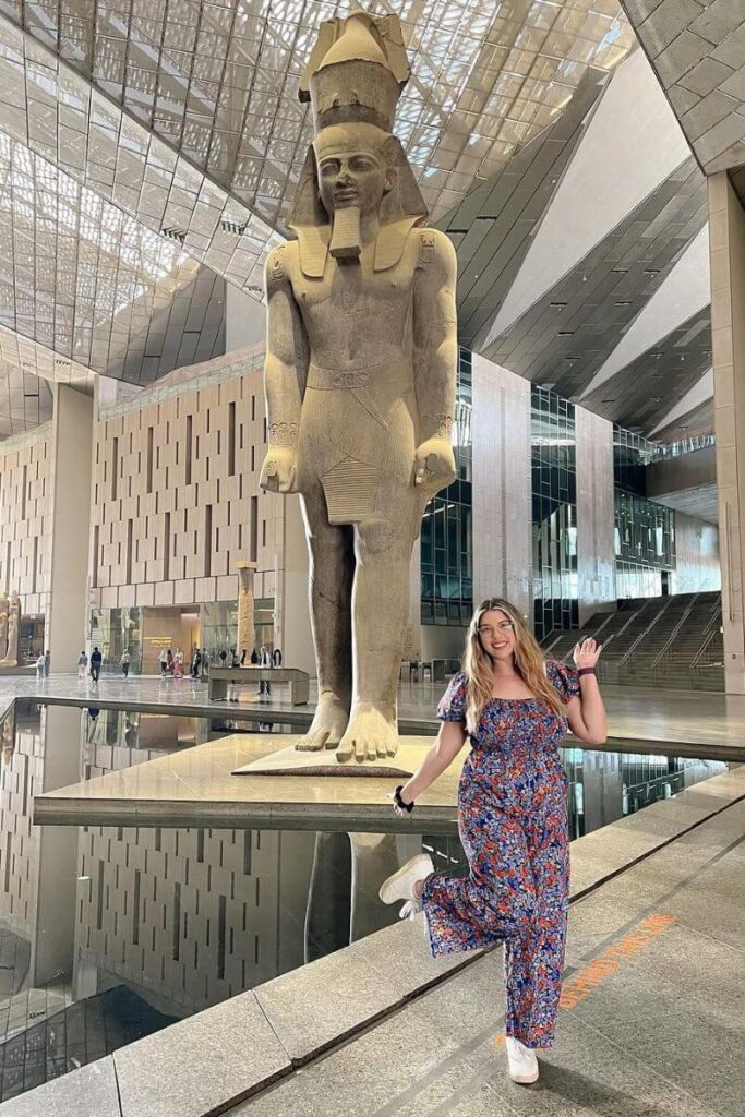 Tourist posing in front of a towering ancient statue in the sleek, modern interior of the Grand Egyptian Museum. A visit here is a must-do in Egypt and among the most exciting new Egypt experiences for culture lovers.