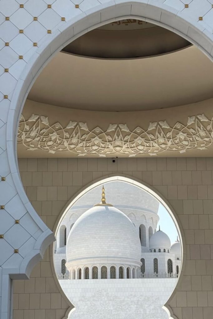 Ornate white domes and gold accents of the Sheikh Zayed Grand Mosque are framed by a decorative arch, glowing in natural light. A top cultural spot in Abu Dhabi, this is one of the best destinations in October for travelers seeking warm weather and architectural beauty.
