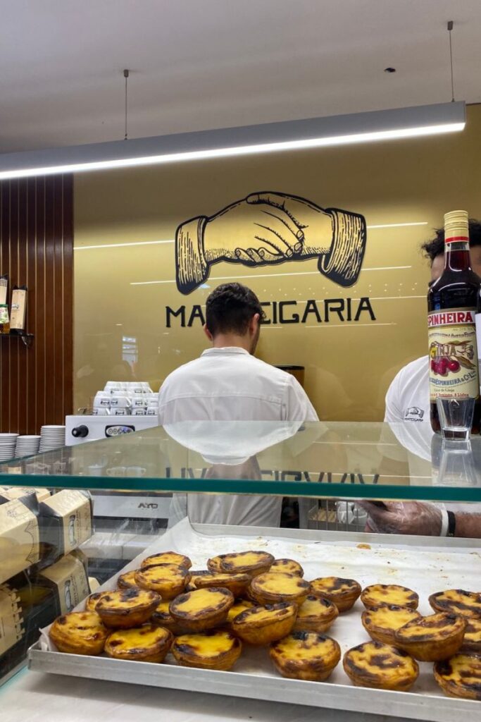 A tray of freshly baked pastel de nata custard tarts sits at the counter of a Lisbon bakery, with staff preparing orders behind glass. Portugal is one of the best warm European destinations in October, offering sunny weather and delicious food culture.