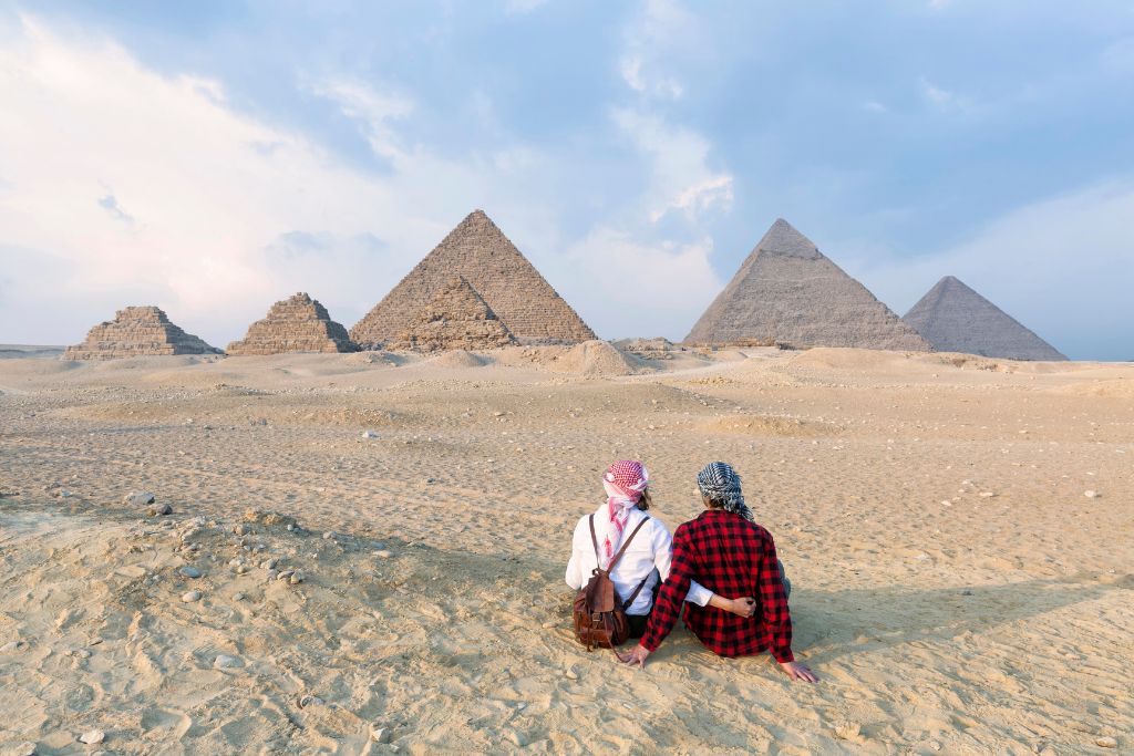 A couple dressed in traditional scarves sits in the desert gazing at the Great Pyramids of Giza under a soft sky. Egypt offers one of the warmest places in October for honeymoon destinations or adventurous vacations filled with history.