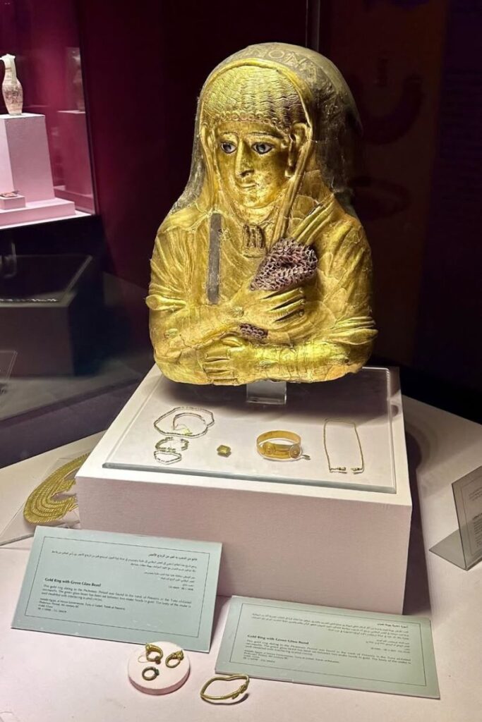 A gold-gilded Greco-Roman mummy mask surrounded by ancient jewelry on display at a museum. Visiting historical sites like this is one of the best day trips from Hurghada and a meaningful way to combine learning with leisure in your list of Hurghada activities.