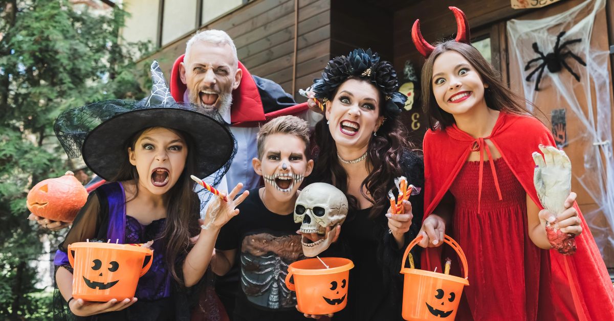 A family dressed as a witch, vampire, skeleton, gothic queen, and devil smiles while holding pumpkin candy buckets, capturing where to go for Halloween and places to visit for Halloween for fun, family-friendly celebrations.