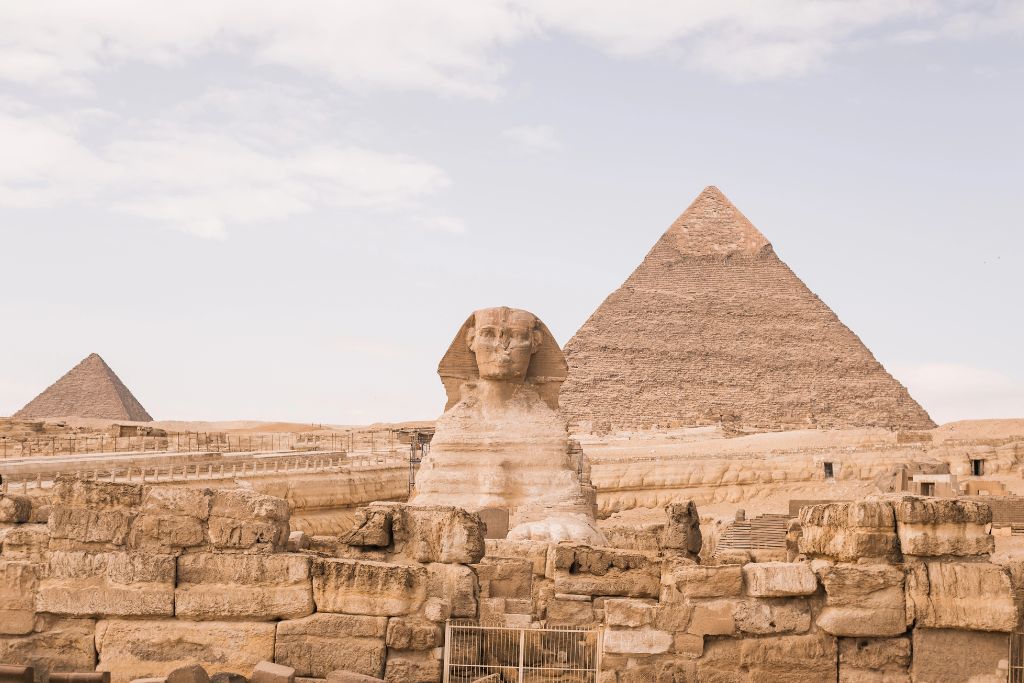 The Great Sphinx in front of the iconic pyramids of Giza under a pale sky, surrounded by ancient ruins and sandstone blocks. Visiting this world wonder is a popular option for day trips from Hurghada and gives insight into history for those traveling to Cairo from Hurghada.