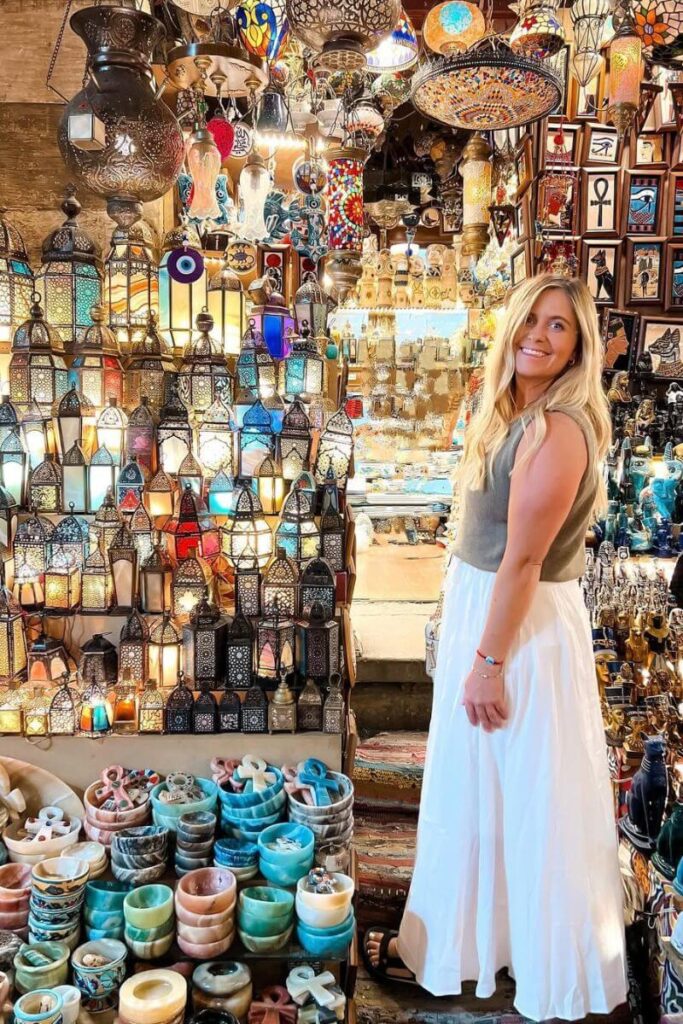 A woman in a white skirt and tank top smiles while browsing a colorful market filled with Egyptian lanterns and handmade goods. This captures Egypt clothing women wear when shopping and offers inspiration for what to wear in Egypt as a woman.